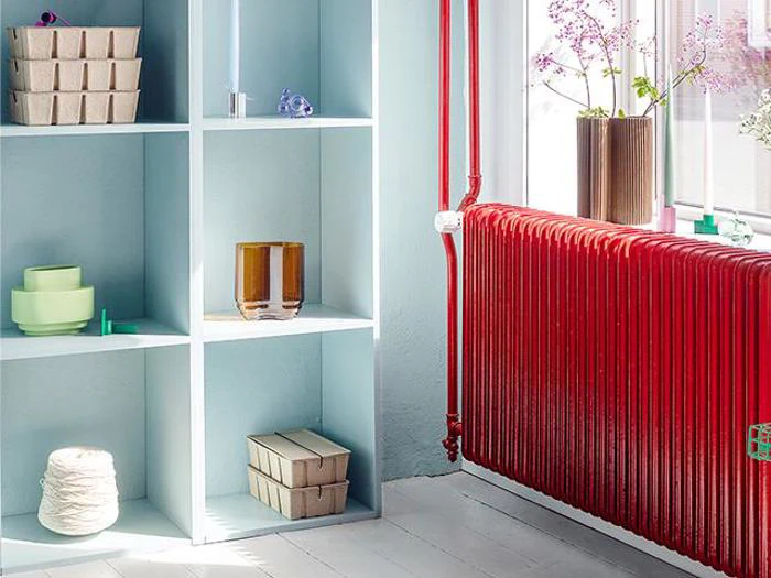 En hvid reol med forskellige pyntegenstande ved siden af en knaldrød radiator under et solbeskinnet vindue med potteplanter.