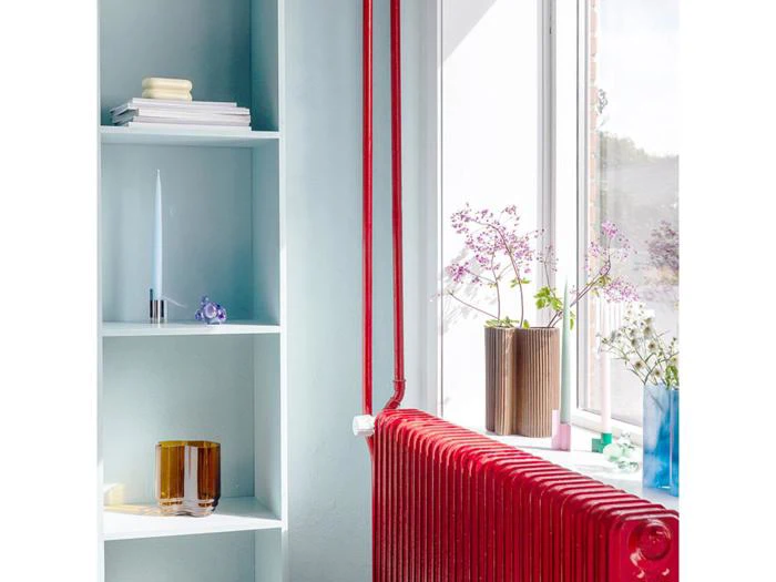 Sollyset strømmer ind gennem et vindue på en rød radiator, glasvaser med blomster og nærliggende lyseblå hylder med bøger og pyntegenstande.