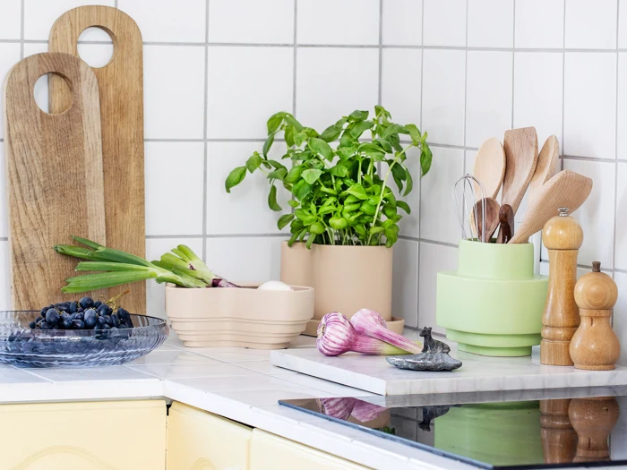 Et ryddeligt køkkenbord med skærebrætter, frisk basilikum, køkkenredskaber, en skål med vindruer, forårsløg, hvidløg og salt- og peberkværne mod et flisebelagt stænk.
