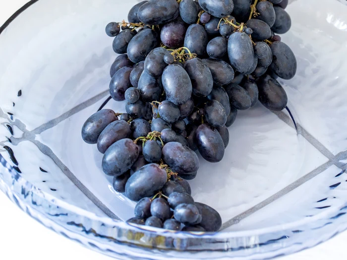 En klase mørklilla vindruer hviler i en klar glasskål med et geometrisk mønster i bunden.