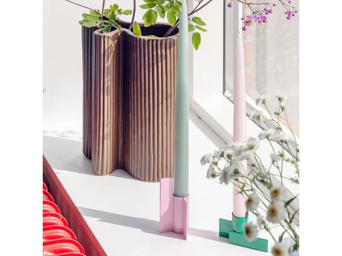 To pastelfarvede stearinlys i moderne stager står på en hvid flade ved et vindue, med en ribbet brun vase med grønt og lilla blomster i baggrunden. Tusindfryd er i forgrunden.