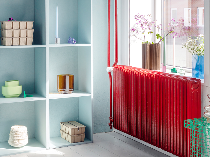 Et lyst rum med en rød radiator under et vindue, åbne hylder med pastelfarvet keramik og æsker og blomster i vaser i vindueskarmen.