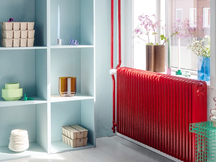 Et lyst rum med en rød radiator under et vindue, åbne hylder med pastelfarvet keramik og æsker og blomster i vaser i vindueskarmen.