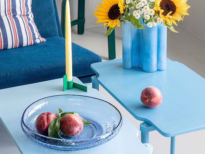 På et blåt bord står en glastallerken med to ferskner, et gult stearinlys i en holder og endnu en fersken i nærheden. I baggrunden står en vase med solsikker og hvide blomster.