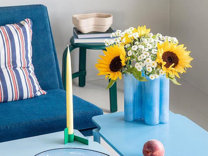 En blå vase med solsikker og hvide margueritter står på et lyseblåt bord ved siden af en stribet pude, et stearinlys og en fersken i en moderne, farverig stue.