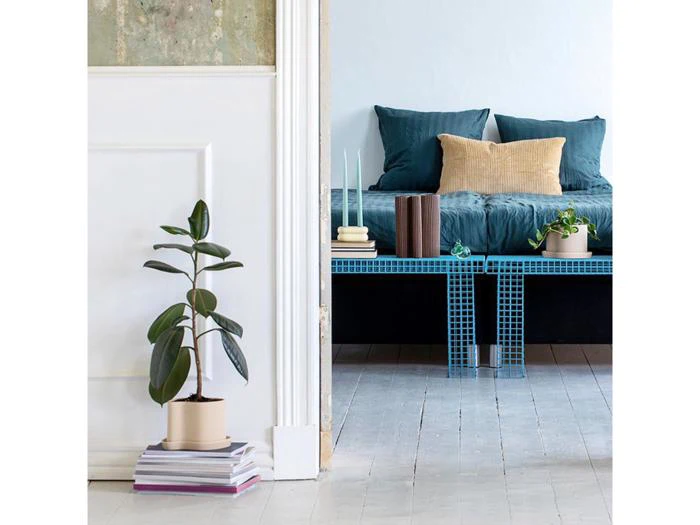 En potteplante står på stablede bøger ved siden af en døråbning, som afslører en seng med blåt sengetøj, en beige pude og to planter på en blå bænk med fliser i et lyst, minimalistisk rum.