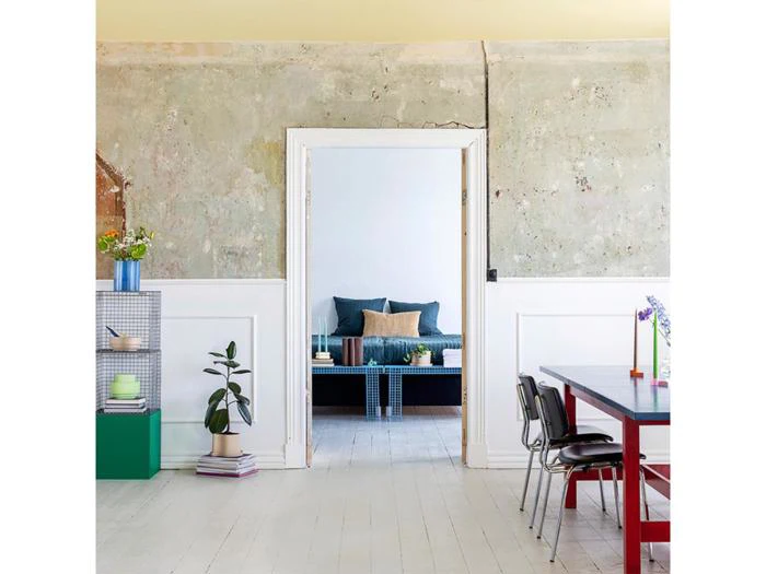 En moderne spiseplads med et rødt bord, sorte stole, stueplanter, åbne reoler og udsigt gennem en døråbning til en hyggelig stue med en blå sofa og minimalistisk indretning.