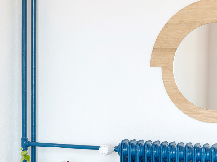 En blå radiator med matchende blå rør under et rundt spejl med træramme, alt sammen opstillet mod en hvid væg.