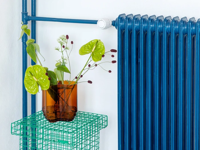 En brun glasvase med grønne og vinrøde blomster står på et grønt trådbord ved siden af en lyseblå radiator på en hvid væg.