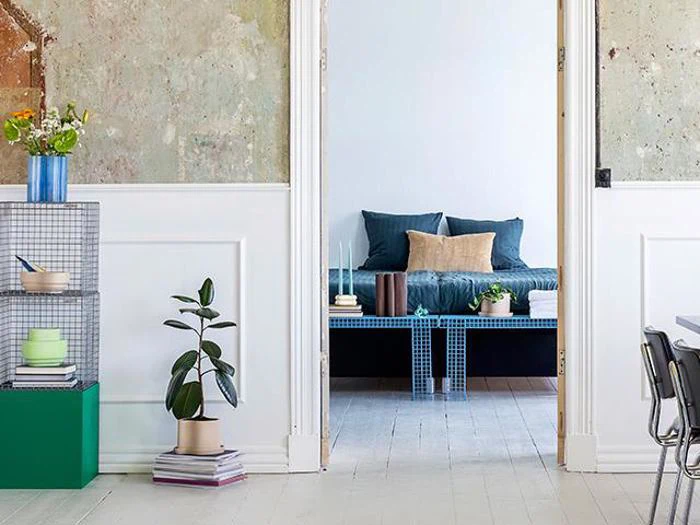 Et moderne opholdsrum med en blå trådbænk, potteplanter, bøger og en hyggelig seng med blå og beige puder i baggrunden.