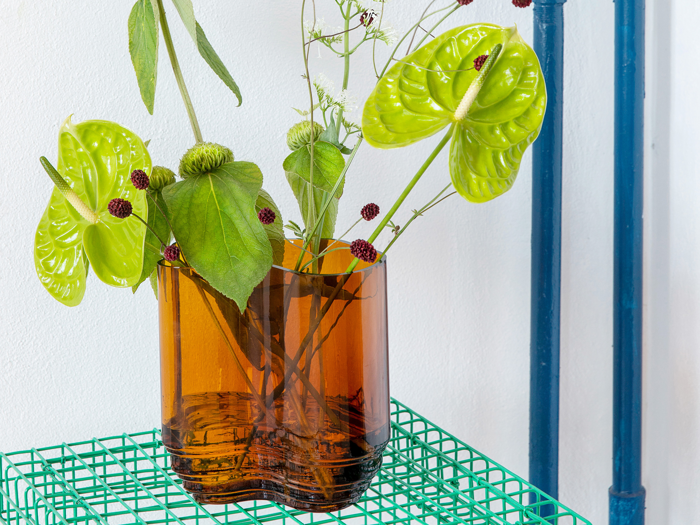 En brun glasvase med grønne anthuriumblomster og blade står på en grøn trådhylde ved siden af blå rør og en hvid væg.