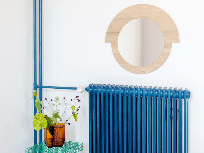 Et rundt træspejl hænger over en blå radiator. På en grøn trådhylde står en ravfarvet glasvase med grønne blade og små hvide blomster.