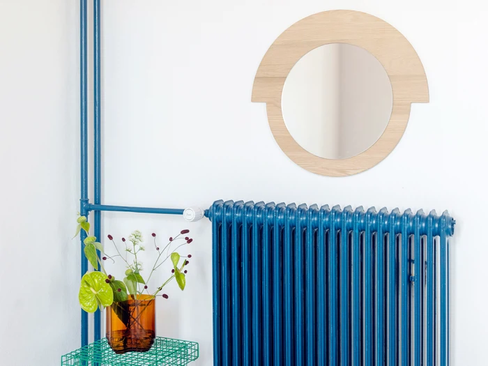 Et rundt træspejl hænger over en blå radiator. På en grøn trådhylde står en ravfarvet glasvase med grønne blade og små hvide blomster.