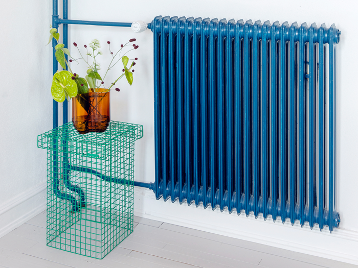En høj blå radiator med forbundne rør står ved siden af et grønt trådbord med en ravfarvet vase med grønne planter, alt sammen mod en hvid væg og et lyst trægulv.