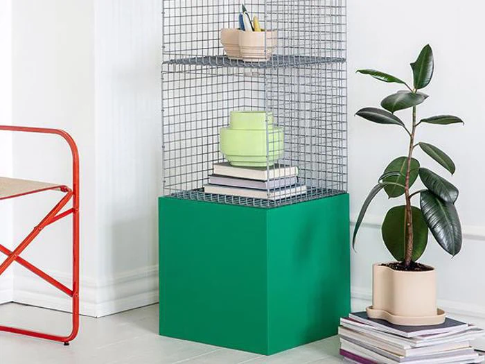 En metaltrådshylde med bøger og dekorationer står på en grøn kube ved siden af en gummiplante i en beige potte og stablede bøger i nærheden af en rød stol mod en hvid væg.