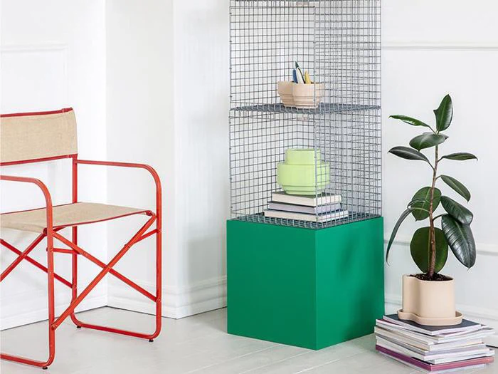 Et moderne interiør med en lærredsstol med rød ramme, en trådhylde på en grøn base med bøger og indretning og en potteplante ved siden af stablede bøger.