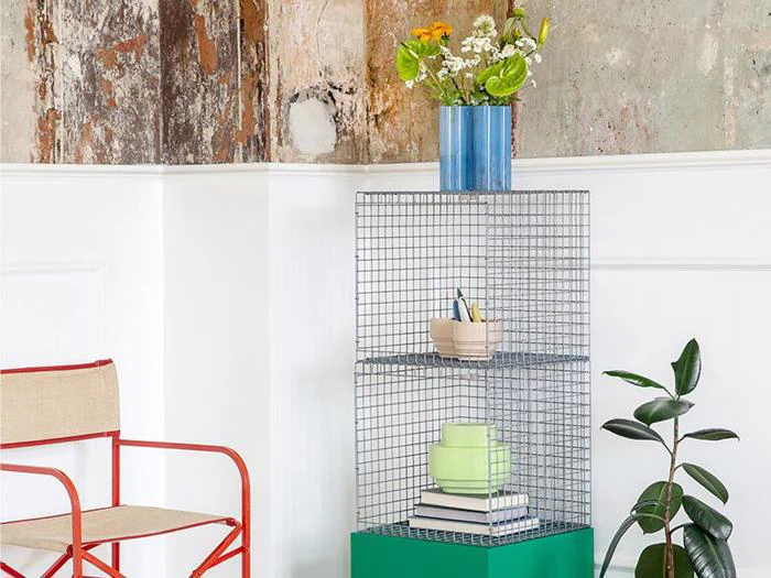 En trådreol med bøger, en blå vase med blomster på toppen, en potteplante og en beige stol med rødt stel ved siden af en struktureret væg.