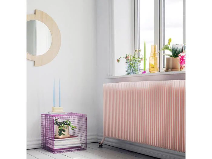 Et lyst rum med en pink radiator, et vindue med farverige vaser og planter, et rundt spejl og et lilla trådbord med bøger, stearinlys og en lille plante.