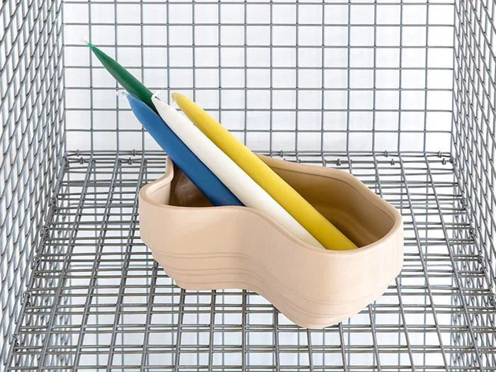 En beige, bølget keramikholder med fire lange lys - grønne, blå, gule og hvide - står på en hylde af metaltråd.