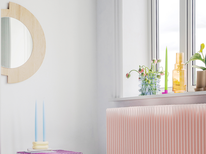 Et lyst rum med et rundt træspejl, en pink radiator, et bord med to blå stearinlys og en vindueskarm med planter og flasker.