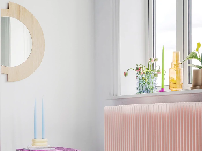 Et lyst rum med et rundt træspejl, en pink radiator, et bord med to blå stearinlys og en vindueskarm med planter og flasker.