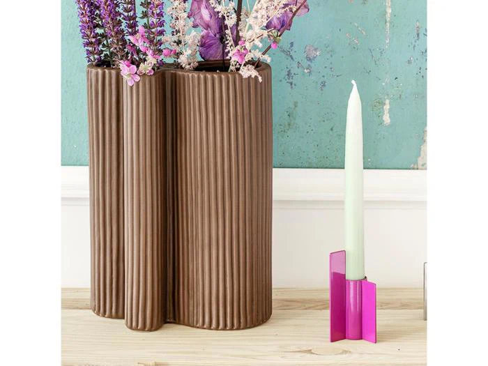 En riflet brun vase med lilla og pink blomster står ved siden af et hvidt lys i en lyserød holder på en træplade mod en slidt turkis væg.
