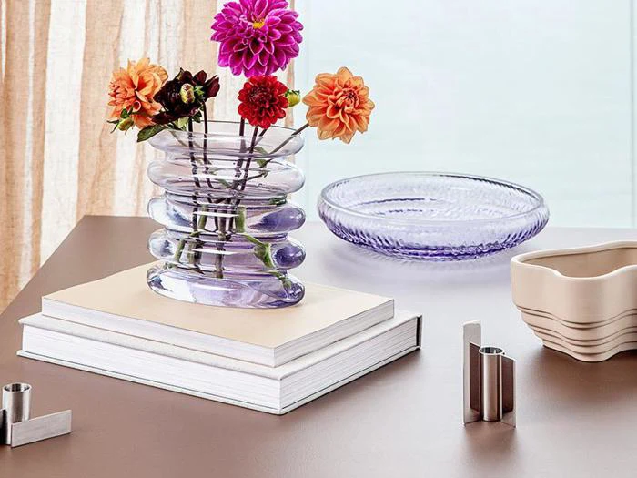 En glasvase med farverige blomster står på en stak bøger ved siden af et lilla glasfad og en beige, bølget skål på et brunt bord.