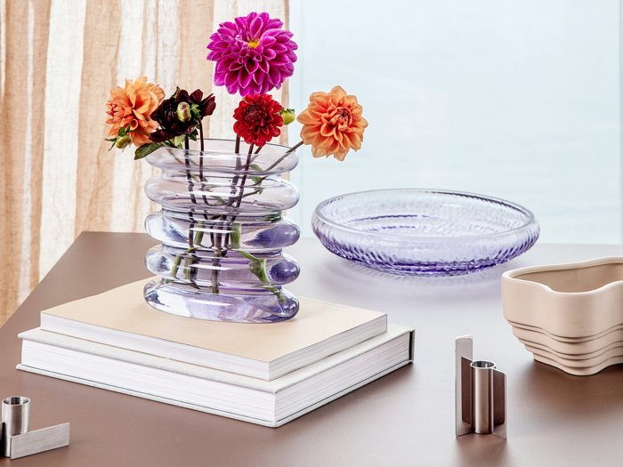 En bølget glasvase med farverige georginer står på stablede bøger ved siden af et lilla glasfad og en beige skål på et lyst træbord ved et vindue.