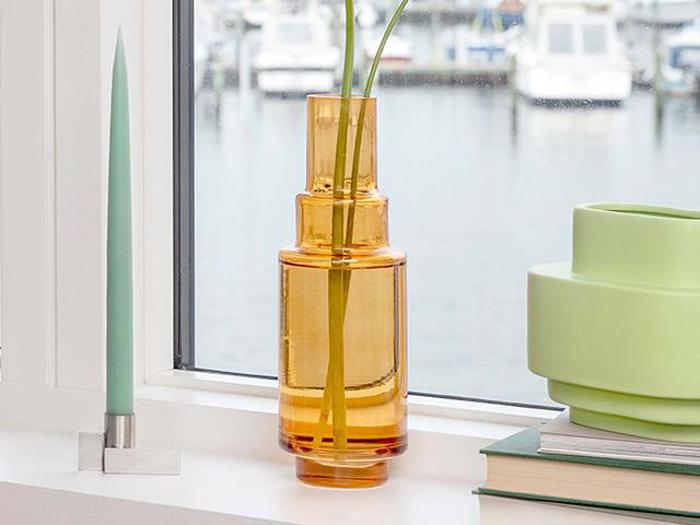 En gul-orange glasvase med lange grønne stilke står i vindueskarmen ved siden af et mintgrønt stearinlys og en stak bøger. Uden for vinduet ses en sløret marina med både.