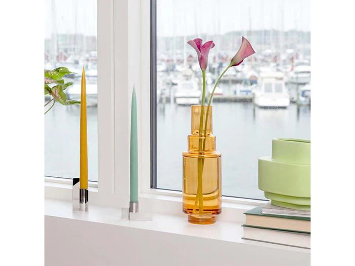 En vindueskarm med gule og grønne stearinlys, en ravfarvet glasvase med to lilla blomster, stablede lysegrønne skåle og bøger med udsigt til en marina med både.