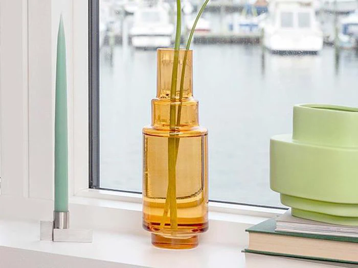 En gul-orange glasvase med lange grønne stilke står i vindueskarmen ved siden af et mintgrønt stearinlys og en stak bøger. Uden for vinduet ses en sløret marina med både.