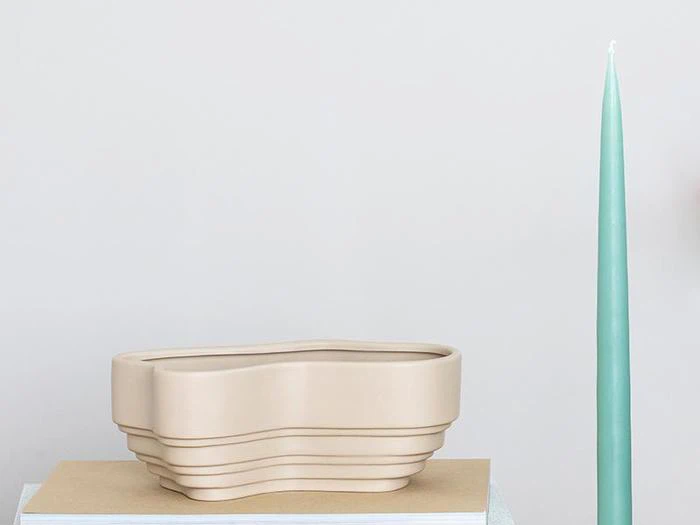 En beige, moderne krukke med bølgede kanter står på en bog ved siden af et højt, slankt lysegrønt lys på en almindelig lys baggrund.