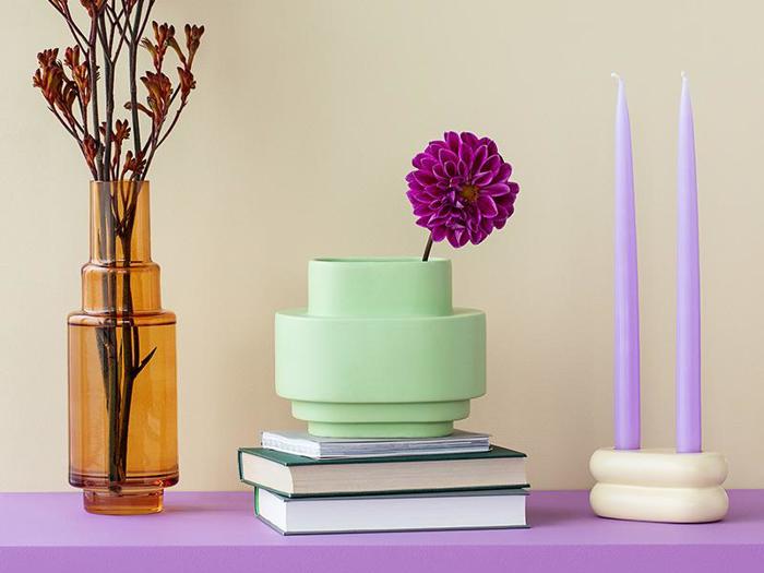 En brun glasvase med grene, en grøn vase med en lilla blomst, stablede bøger og to høje lavendelfarvede stearinlys på en lilla flade mod en beige baggrund.