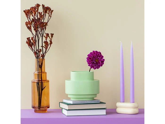 Et stilleben med tørrede blomster i en ravfarvet vase, en lilla blomst i en grøn vase, stablede bøger og to lavendelfarvede stearinlys på en lilla overflade mod en beige baggrund.