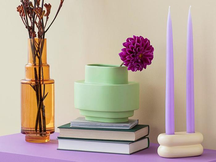 Et moderne stilleben med en ravfarvet vase med tørrede grene, en grøn vase med en lilla blomst, stablede bøger og to høje lavendellys på en lilla flade.