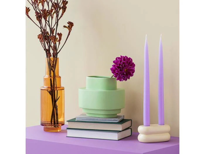 Et stilleben med en brun glasvase med grene, en grøn vase med en lilla blomst, to bøger og to lavendelfarvede stearinlys på en lilla flade mod en beige væg.