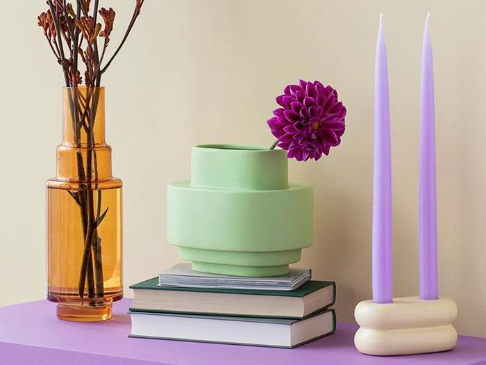 Et moderne stilleben med en ravfarvet vase med tørrede grene, en grøn vase med en lilla blomst, stablede bøger og to høje lavendellys på en lilla flade.