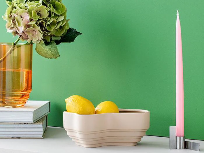 En beige skål med to citroner står på en hvid flade ved siden af et lyserødt lys i en holder, en glasvase med grønne og lyserøde hortensiaer og stablede bøger; grøn væg i baggrunden.