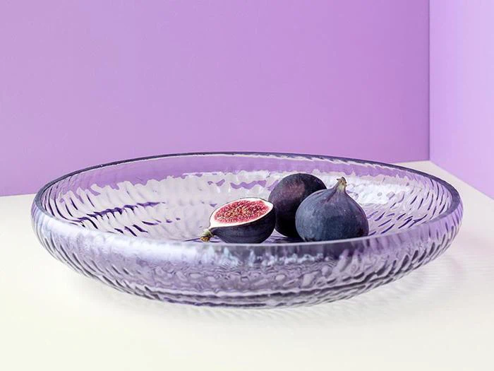 En tekstureret glasskål indeholder tre figner, hvoraf den ene er skåret midt over, på en lyslilla baggrund.