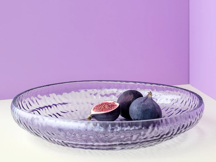 En klar, struktureret glasskål indeholder tre figner, hvoraf den ene er skåret op, på en pastellilla baggrund.