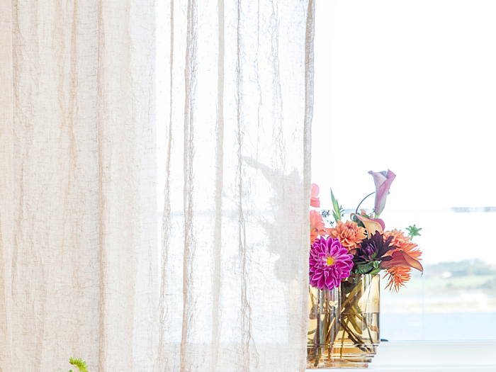 En vase med farverige blomster står i vindueskarmen, delvist skjult bag et gennemsigtigt beigefarvet gardin, med en lys udendørs scene i baggrunden.