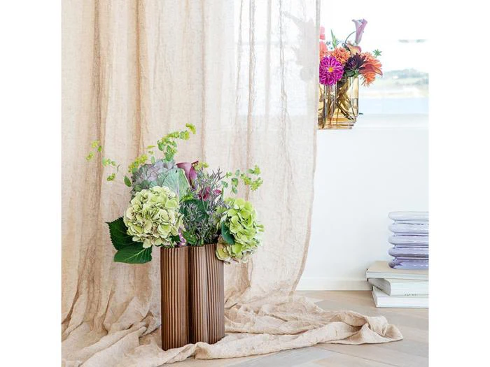 En brun riflet vase med grønne og lilla blomster står på et beige draperet gardin; et vindue og en anden vase med farverige blomster står i baggrunden.