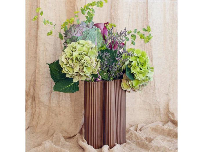 En brun ribbet vase indeholder grønne hortensiaer, lilla blomster og grønne planter på en baggrund af beige stof.