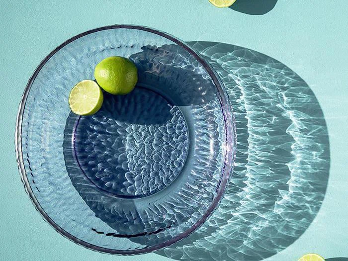 En klar glasskål med to limefrugter indeni kaster mønstrede skygger på en lyseblå overflade med to halve limefrugter i nærheden.