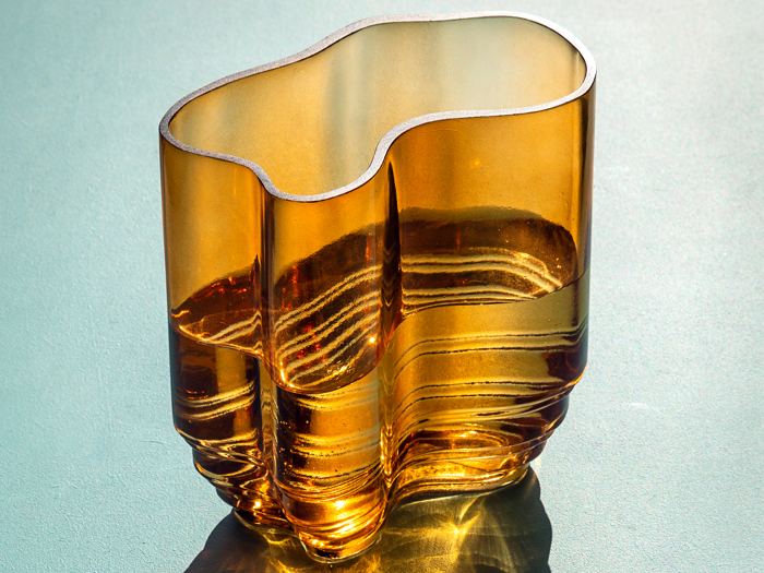 En bølget, ravfarvet glasvase står på en lys flade og kaster komplicerede skygger og refleksioner i sollyset.