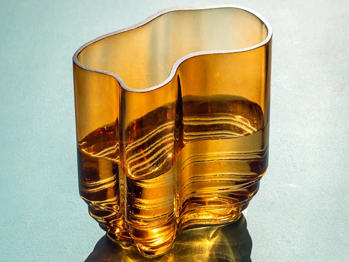 En bølget, ravfarvet glasvase står på en lys flade og kaster komplicerede skygger og refleksioner i sollyset.