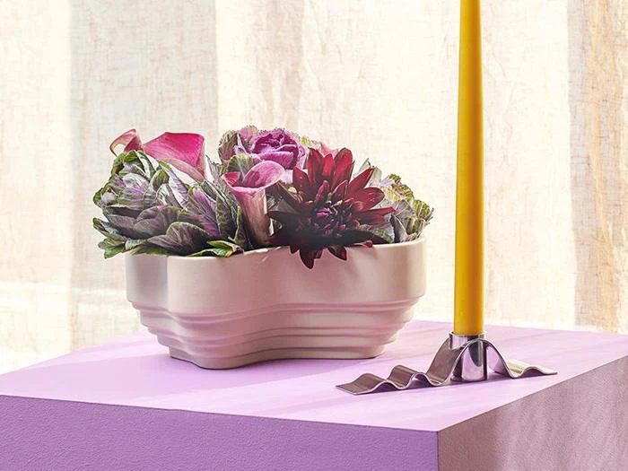 En keramisk krukke med prydkål og blomster står på en lilla flade ved siden af et højt gult lys i en bølget metalholder.