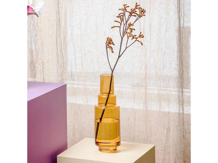 En høj orange glasvase med geometriske former holder en enkelt tørret gren, der står på en cremefarvet overflade med en lilla blok og gennemsigtige gardiner i baggrunden.