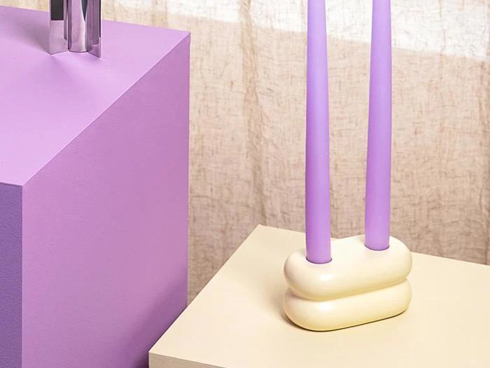 To høje lavendellys i en cremefarvet, afrundet holder står på en lys overflade ved siden af en lilla blok mod et beige tekstureret gardin.