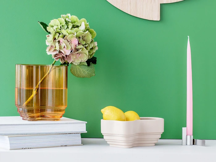 En grøn hortensia i en ravfarvet vase, to citroner i en cremefarvet skål og et højt lyserødt stearinlys på en hvid flade mod en grøn væg.